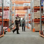 Dua staf gudang memeriksa dan mengelola inventaris barang di lorong gudang penyimpanan